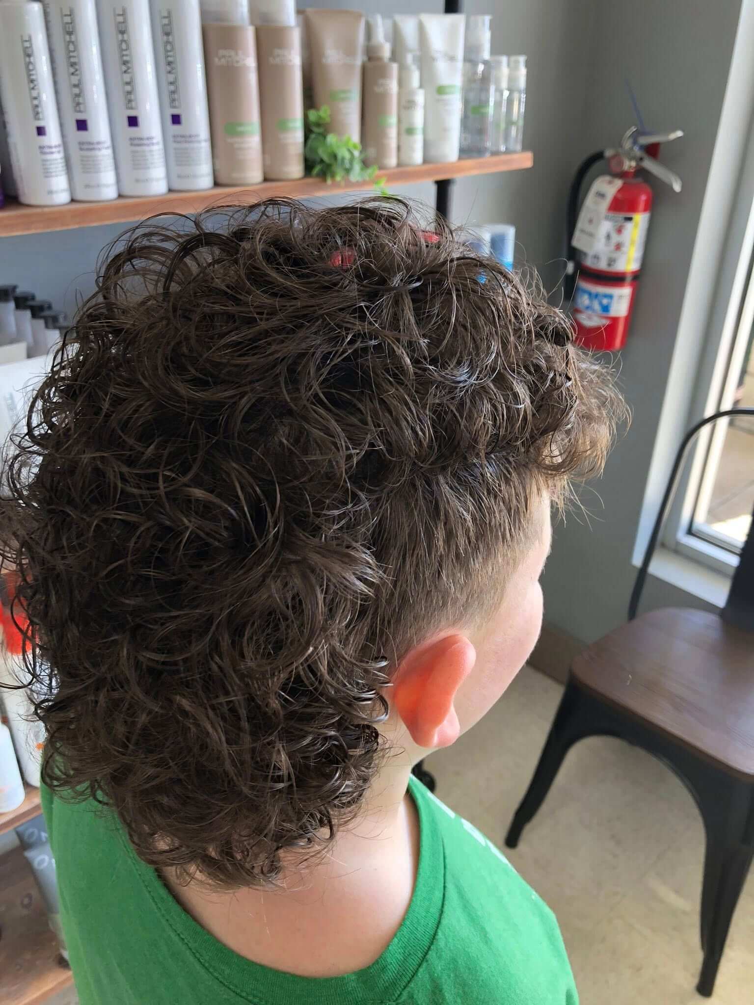 Back and side view of a child with short, curly brown hair wearing a green shirt inside a salon.