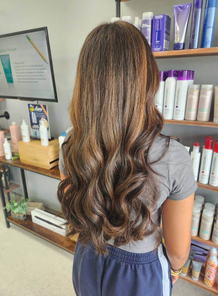 Person with long, wavy brown hair with subtle highlights standing in a salon in front of shelves with hair care products.