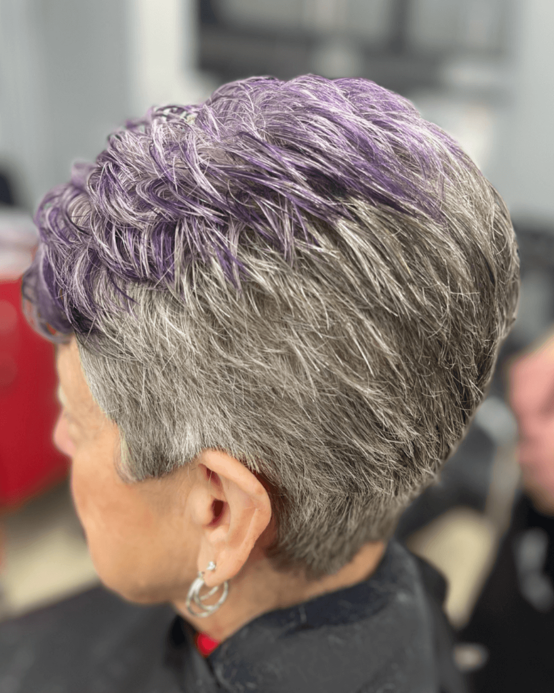 Side view of a person with short gray hair dyed purple on top, wearing silver hoop earrings.