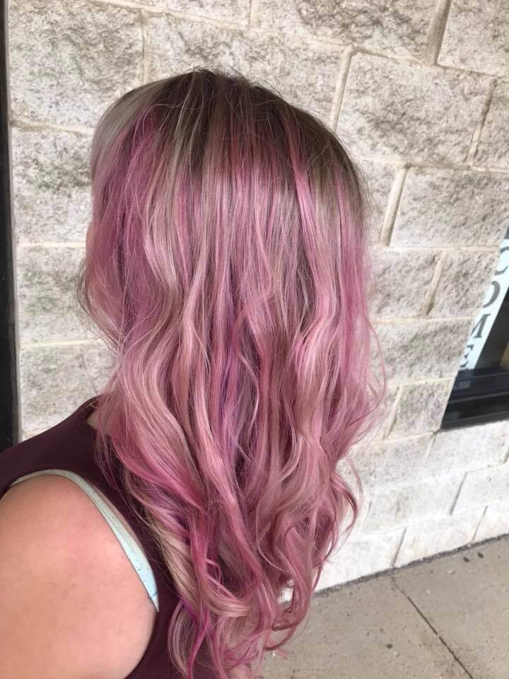Person with long wavy hair dyed in shades of pink and blonde standing against a light stone wall.