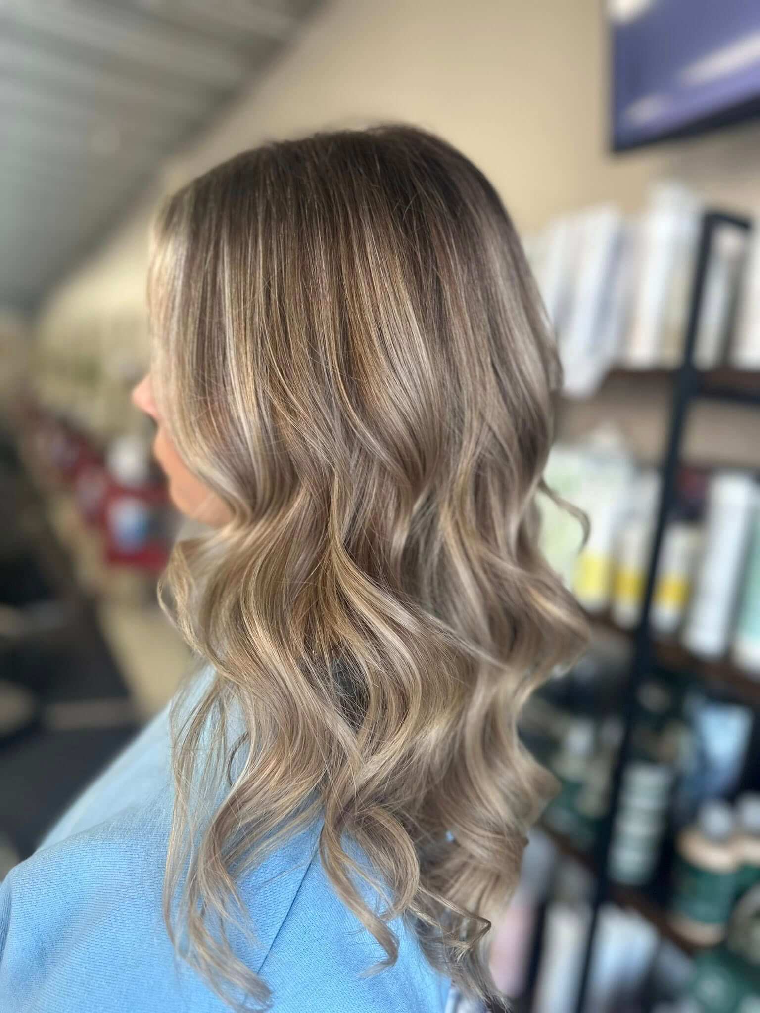 Side view of a person with long, wavy, ash blonde hair wearing a light blue top in a salon setting.