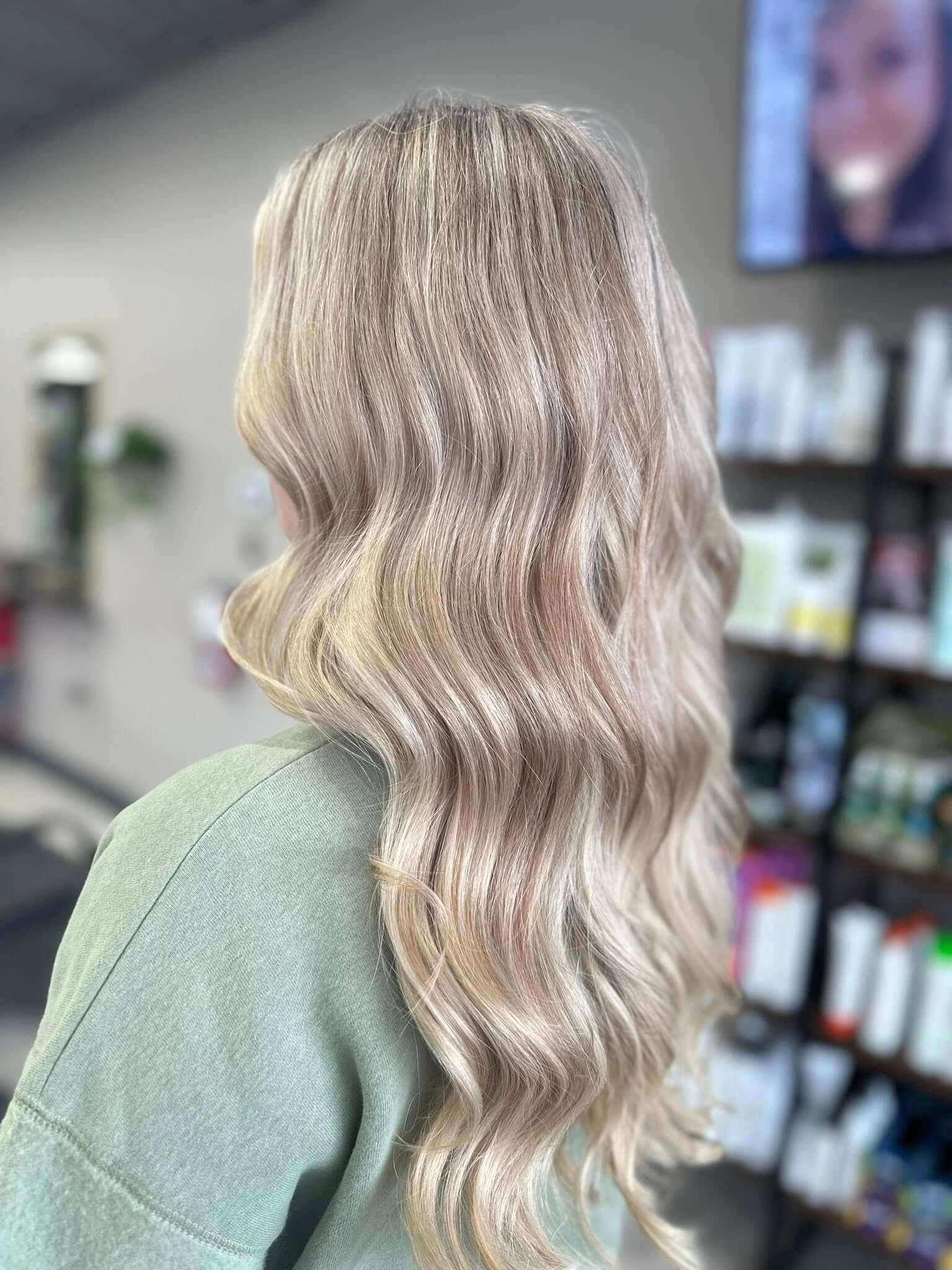 Back view of a person with long, wavy, blonde hair wearing a light green top in a salon setting.