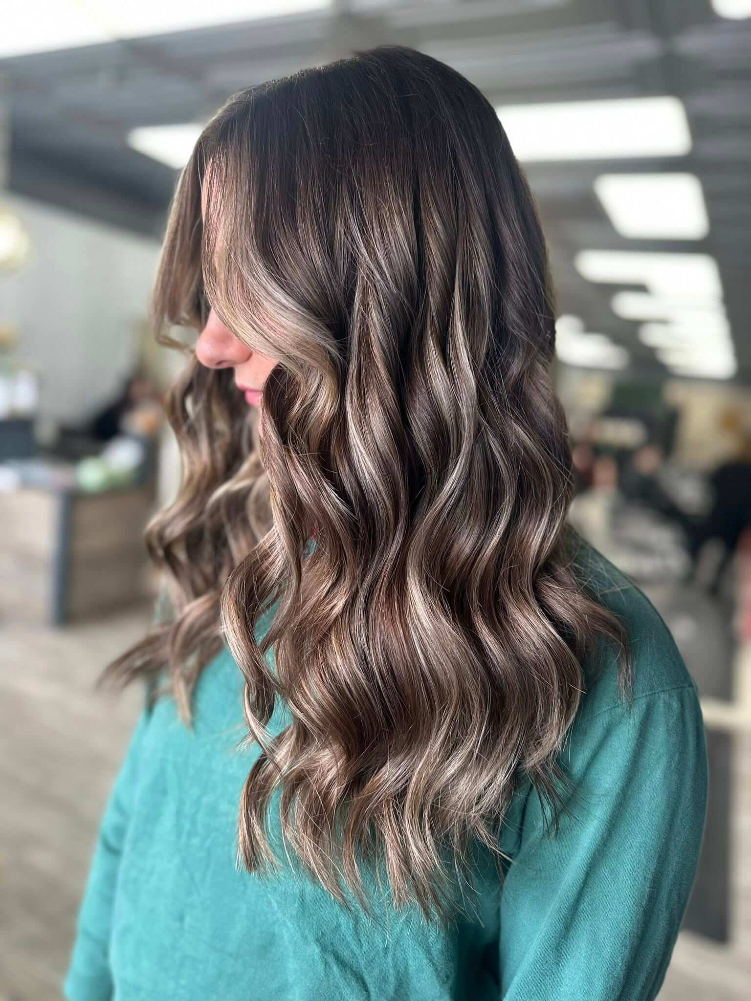 Side view of a woman with long, wavy brown hair with subtle highlights wearing a green top in a salon.