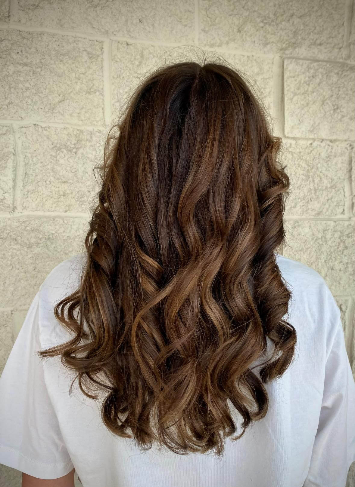 Back view of person with medium-length, wavy brown hair wearing a white t-shirt against a light stone wall.