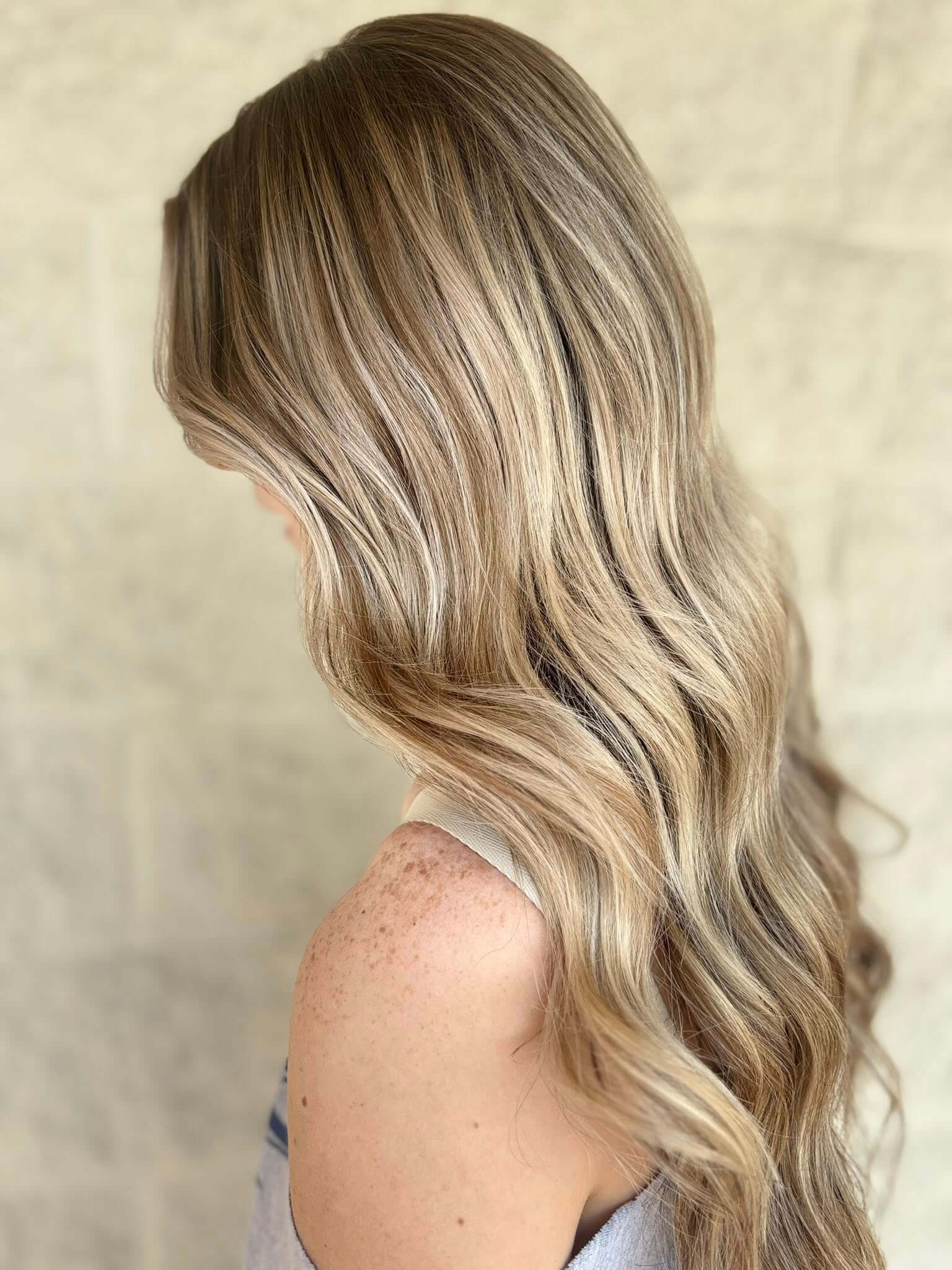 Side view of a person with long, wavy blonde hair and freckles on their shoulder against a neutral background.
