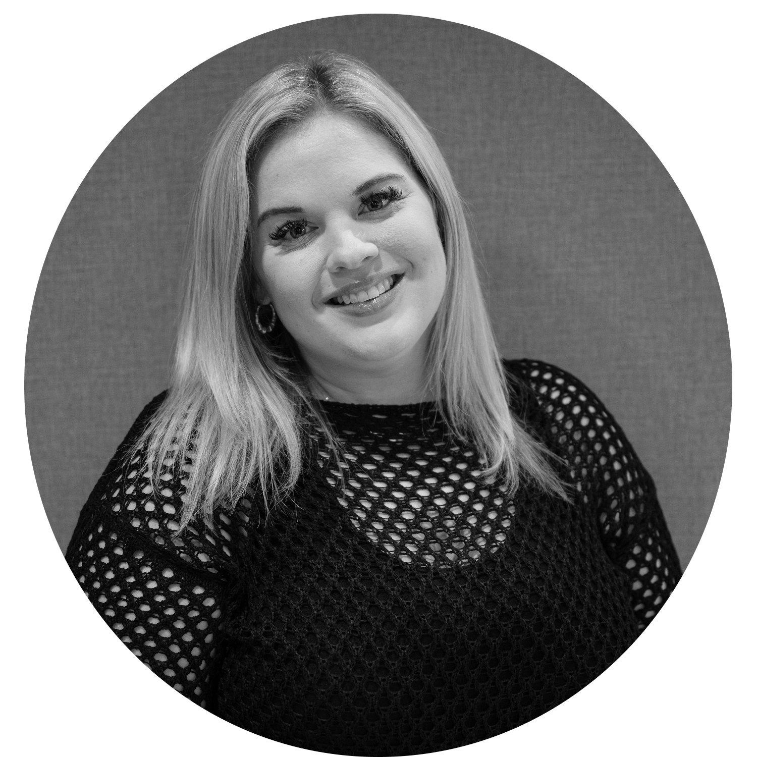 Smiling woman with long wavy hair wearing a black top and layered necklaces standing against a textured wall.