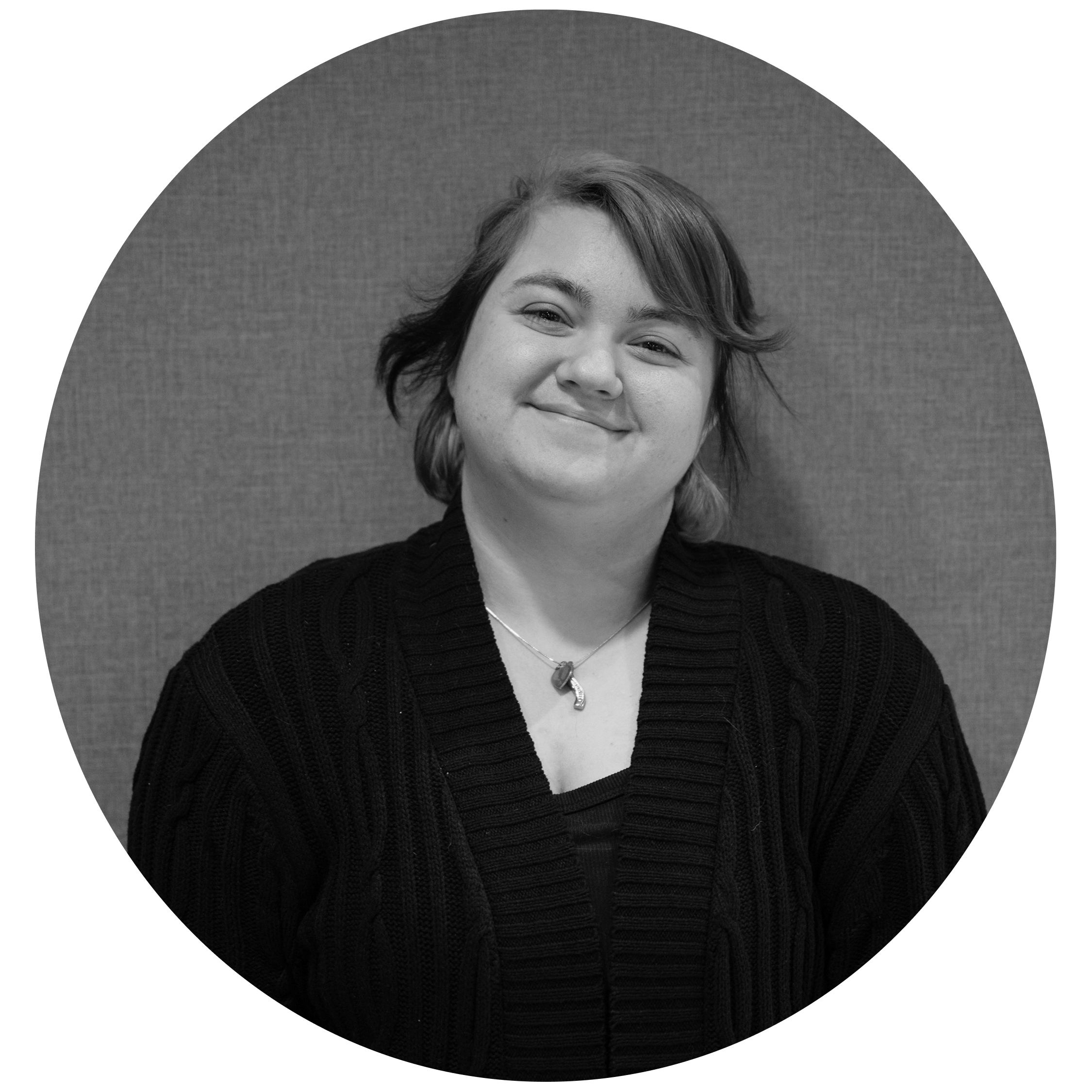 Smiling person with short hair wearing a dark knitted sweater and a necklace, posed against a textured background.