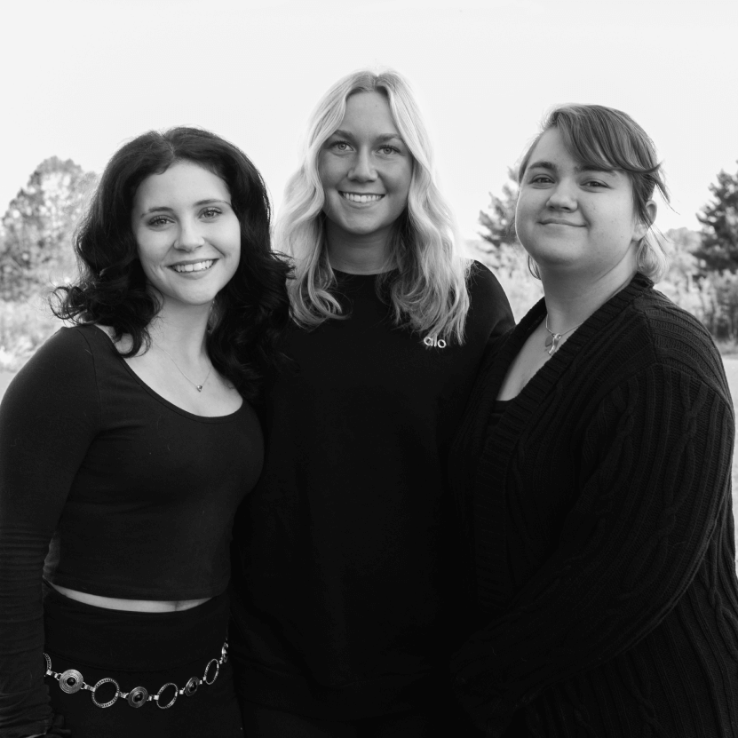 Three women smiling and standing closely together outdoors with trees in the background.