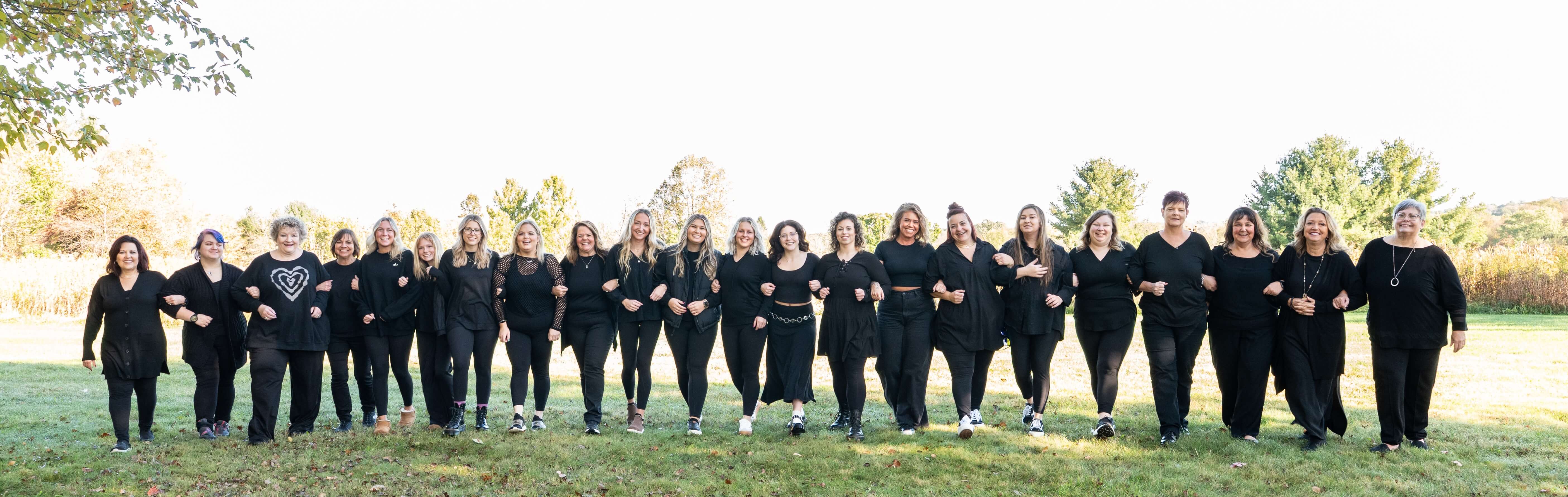 Group of twenty women standing outdoors on grass, linked arm in arm, all wearing black clothing and smiling.