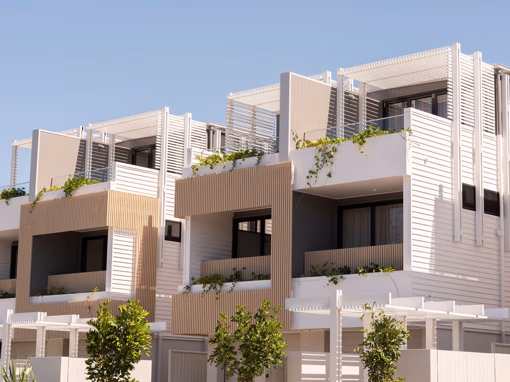 Row of modern two-story townhouses with garages, large windows, trees, and green lawn in front under a blue sky.
