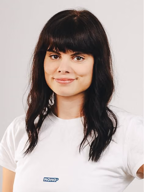 Woman with dark wavy hair and bangs wearing a white NOHO T-shirt against a plain background.