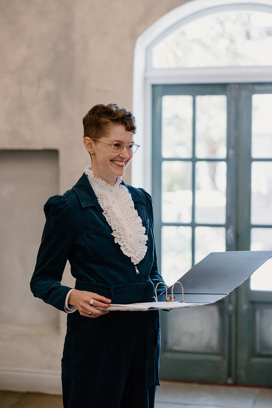 Chloe stands holding a ceremony folder, smiling, in a blue jumpsuit. She's at a wedding, awaiting the arrival of the brides.