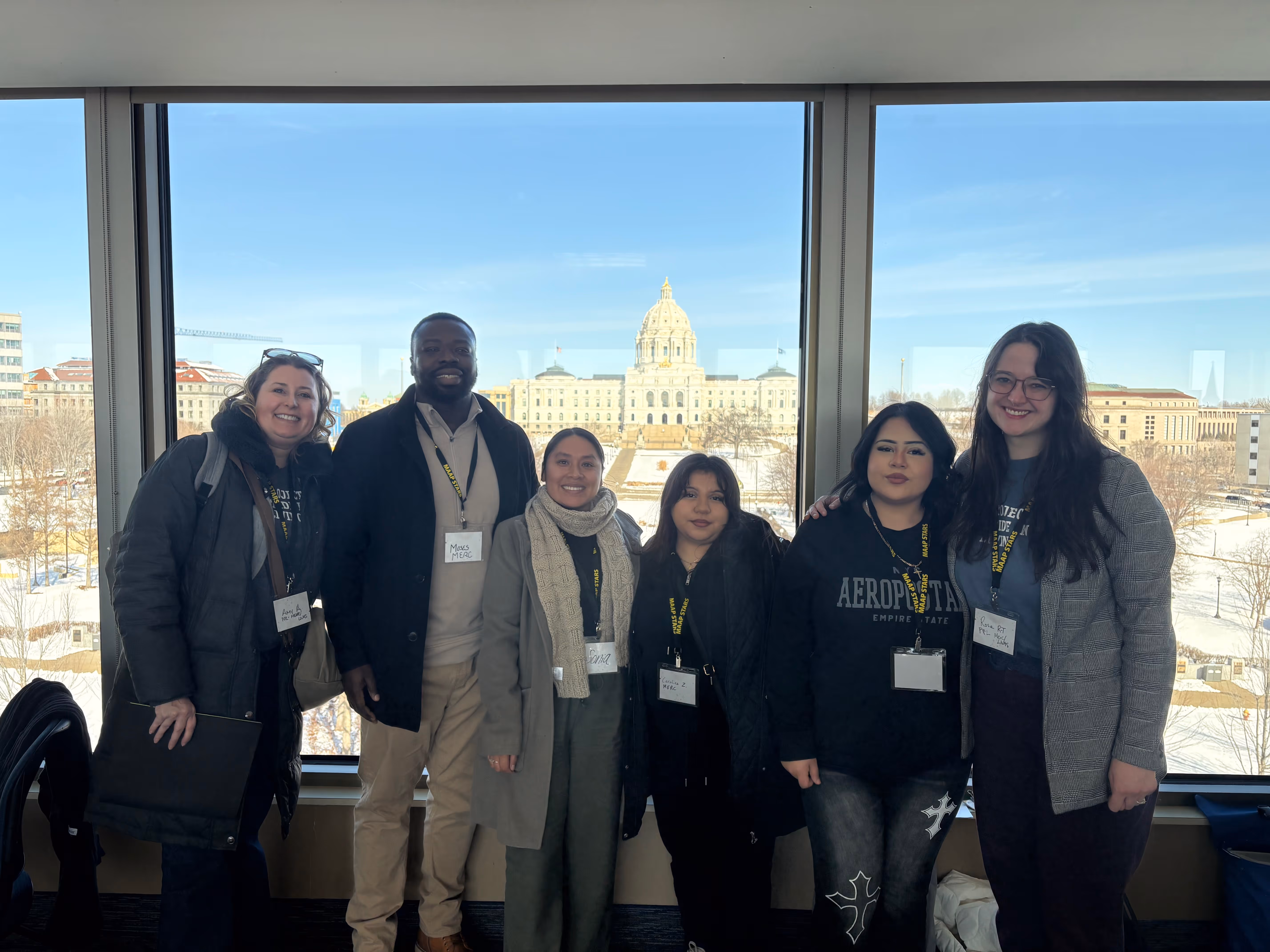 Group of six people smiling and standing indoors in front of large windows showing a snowy urban scene with a domed building outside.
