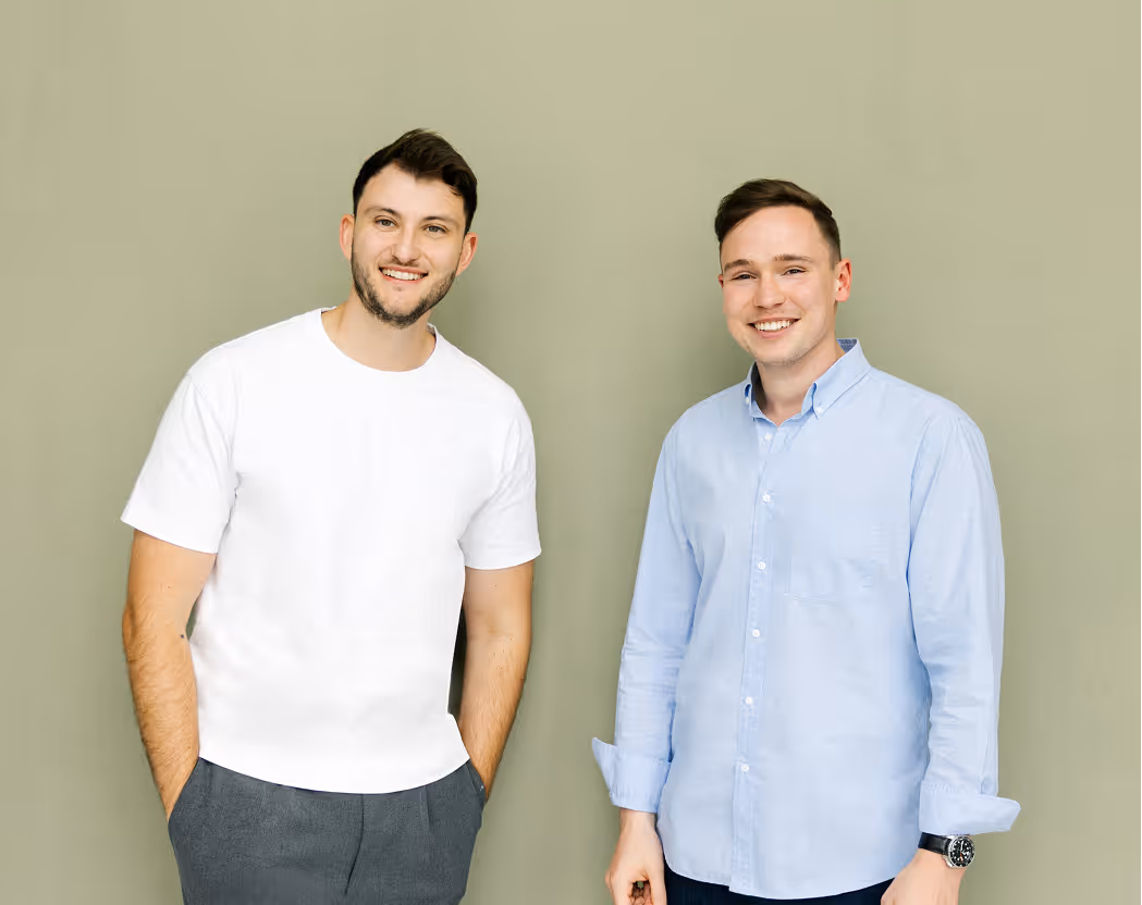 Zwei lächelnde Männer, einer in weißem T-Shirt, der andere in blauem Hemd, vor beigem Hintergrund.