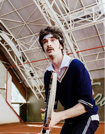 Ein junger mAnn in retro Tennis-Outfit, der in einer Tennishalle steht.