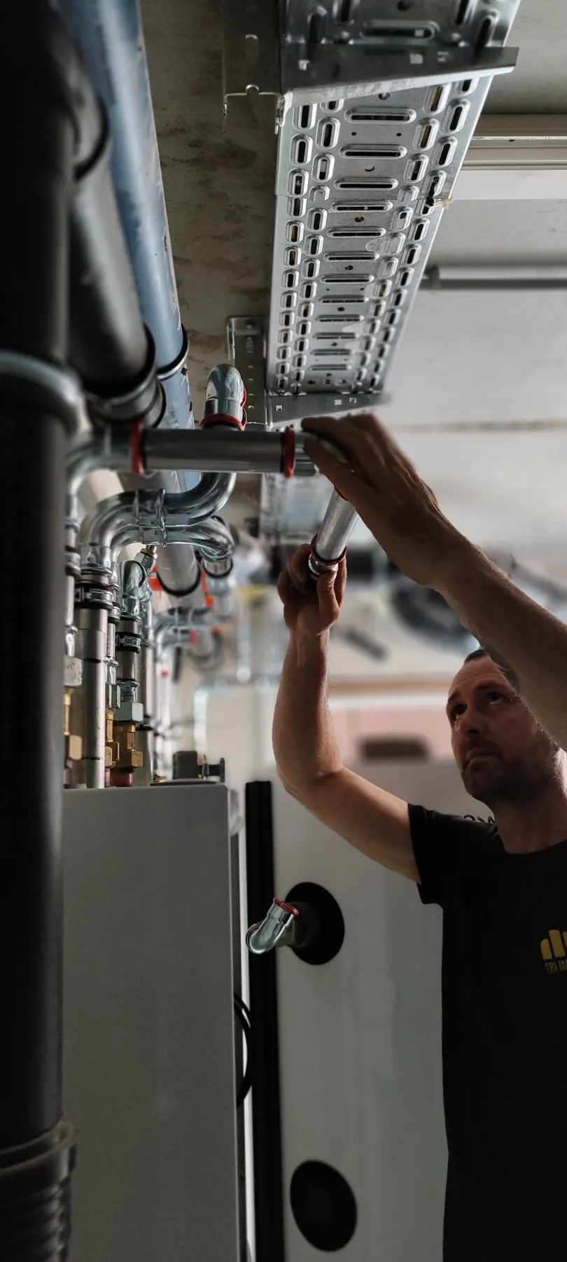 Techniker im TRI-IMMO-Shirt arbeitet an der Verrohrung über einem Speicher im Heizraum