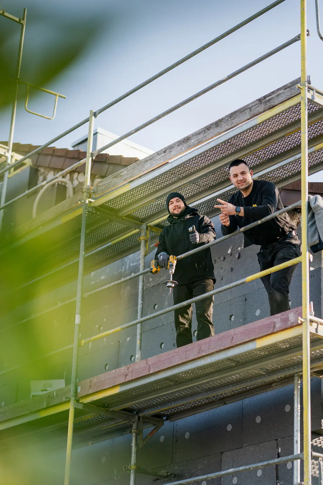 Zwei Mitarbeiter von TRI IMMO stehen auf dem Gerüst einer Fassadensanierung und zeigen nach vorn