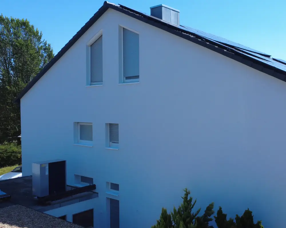 Giebelseite eines Hauses nach der 360° Sanierung: weiße gedämmte Fassade, Solarmodule auf dem Dach, die Wärmepumpe steht auf dem Anbau