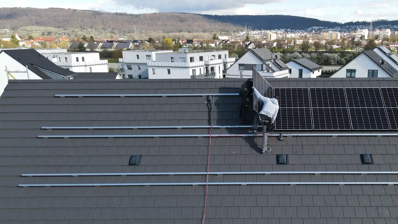 Zwei Monteure setzen von oben gesehen Photovoltaikmodule auf ein dunkles Steildach, daneben die Schienen der Unterkonstruktion