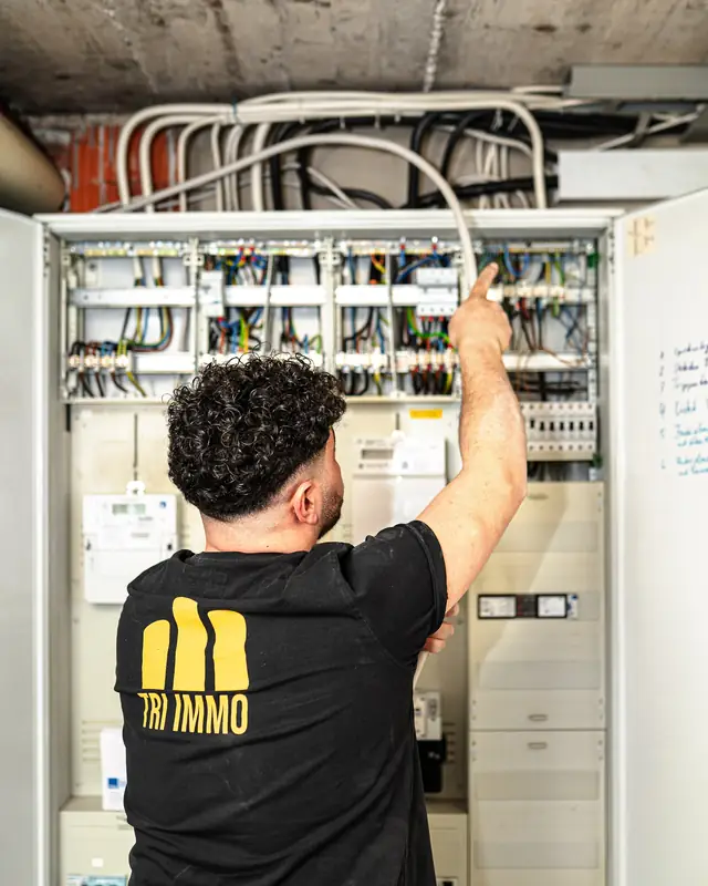 Elektriker verdrahtet einen Zählerschrank, auf dem Rücken das Logo von TRI IMMO