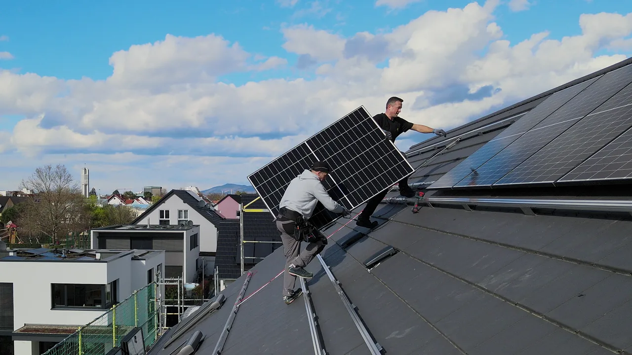 Photovoltaik in Heidelberg und Umgebung: Zwei Monteure tragen ein Photovoltaikmodul über ein dunkles Steildach, von der Drohne aus fotografiert