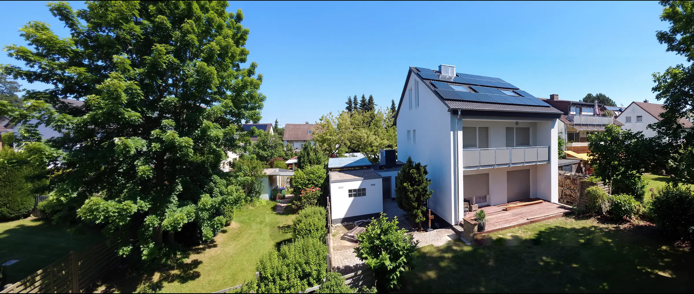 Energetisch saniertes Einfamilienhaus von der Drohne aus: weiße, gedämmte Fassade, neues Dach mit schwarzen Solarmodulen, Balkon mit neuem Geländer, daneben der Garten. Foto, am Rand per KI ergänzt.