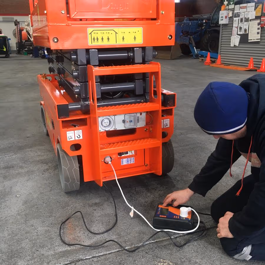 A person is crouching, plugging a scissor lift into a power outlet for charging.
