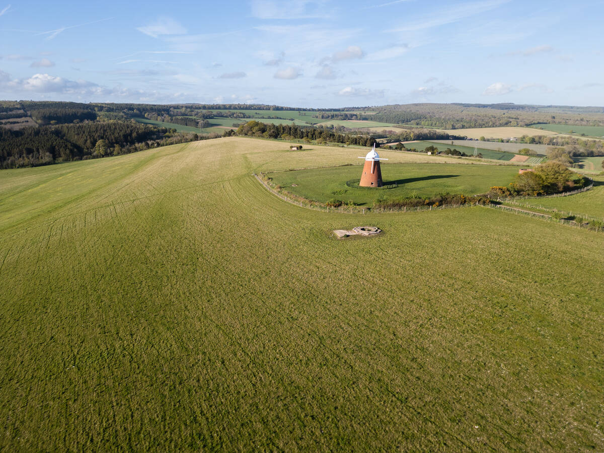 Aerial view of a green hill with a small brick windmill surrounded by a circular fence and countryside in the background under a blue sky.
