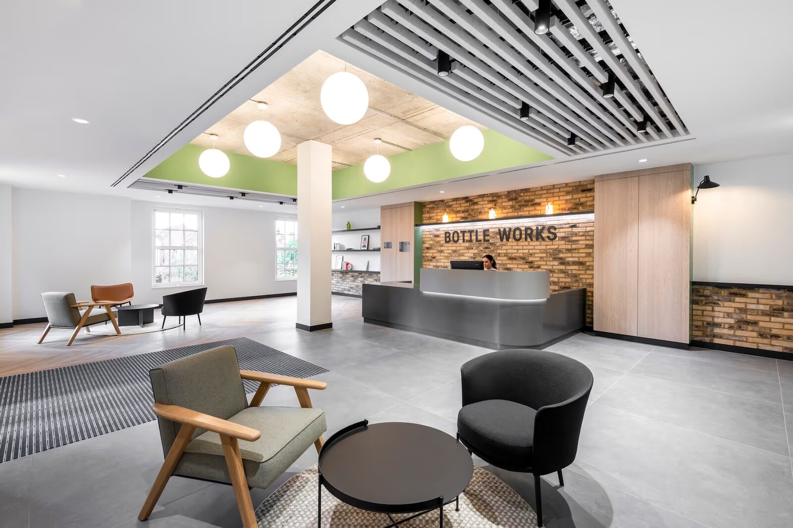 Modern office reception area with a receptionist behind a sleek desk, brick accent wall with 'BOTTLE WORKS' sign, and contemporary seating.