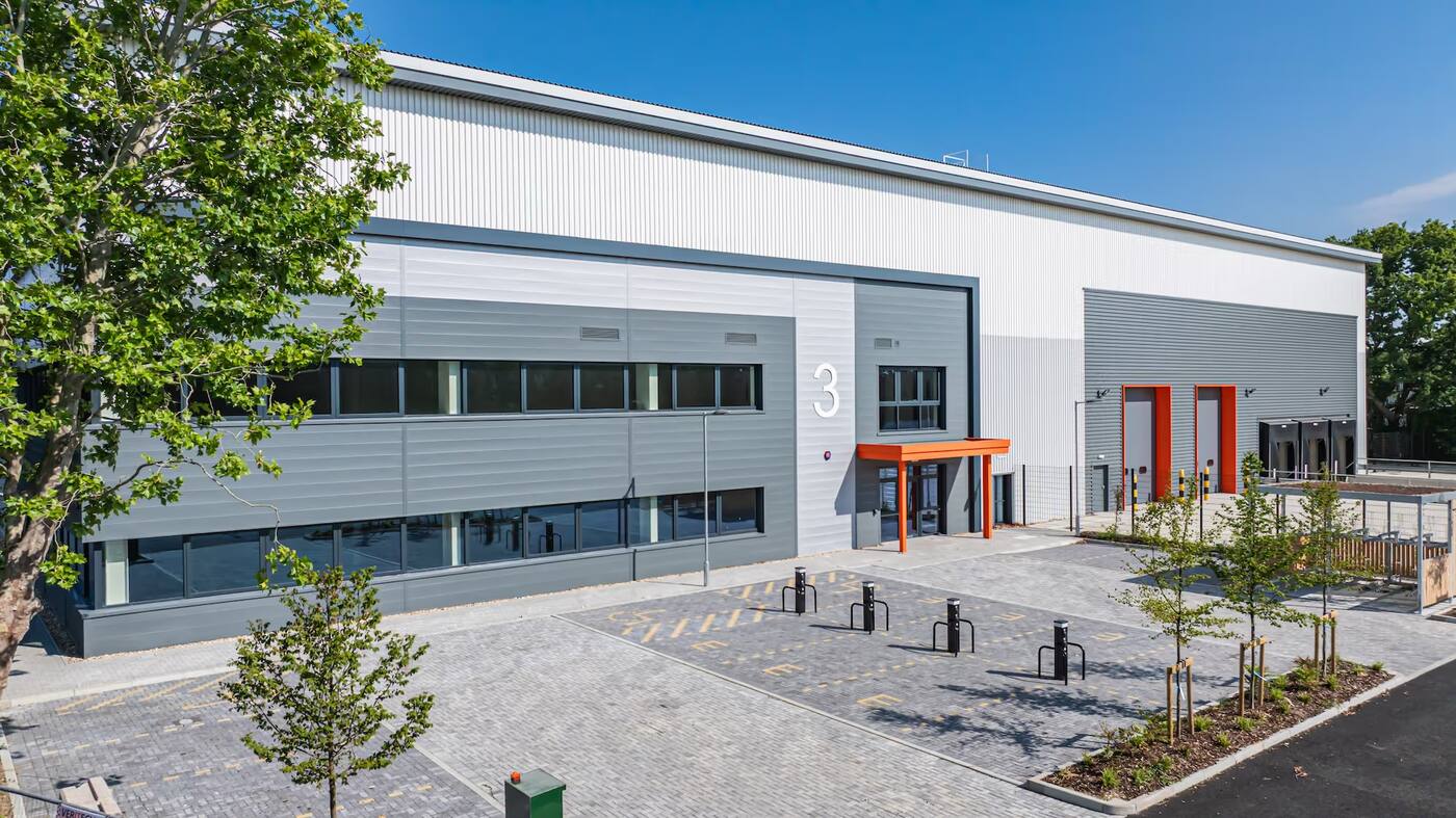 Modern industrial building with gray and white exterior, large number 3, orange entrance canopy, and empty paved parking area with young trees.