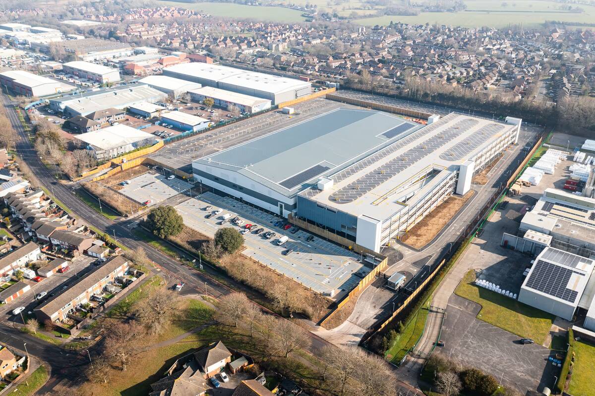 Aerial view of a large industrial warehouse with rooftop solar panels surrounded by parking lots and nearby residential and commercial buildings.