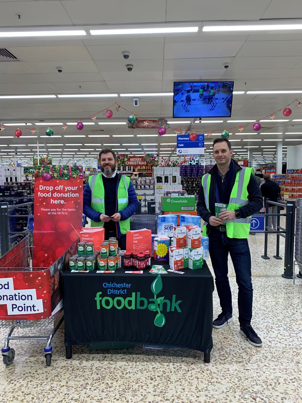 Two men in neon green safety vests standing behind a Chichester District Foodbank donation table inside a supermarket.