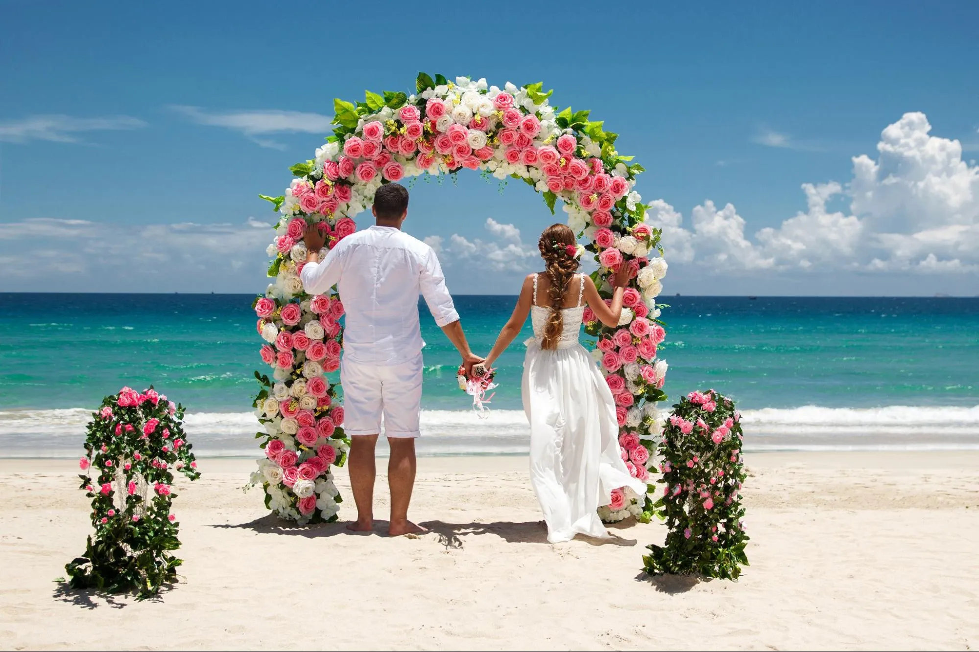 Romantic beach wedding couple holding hands under floral arch with pink and white roses by the ocean