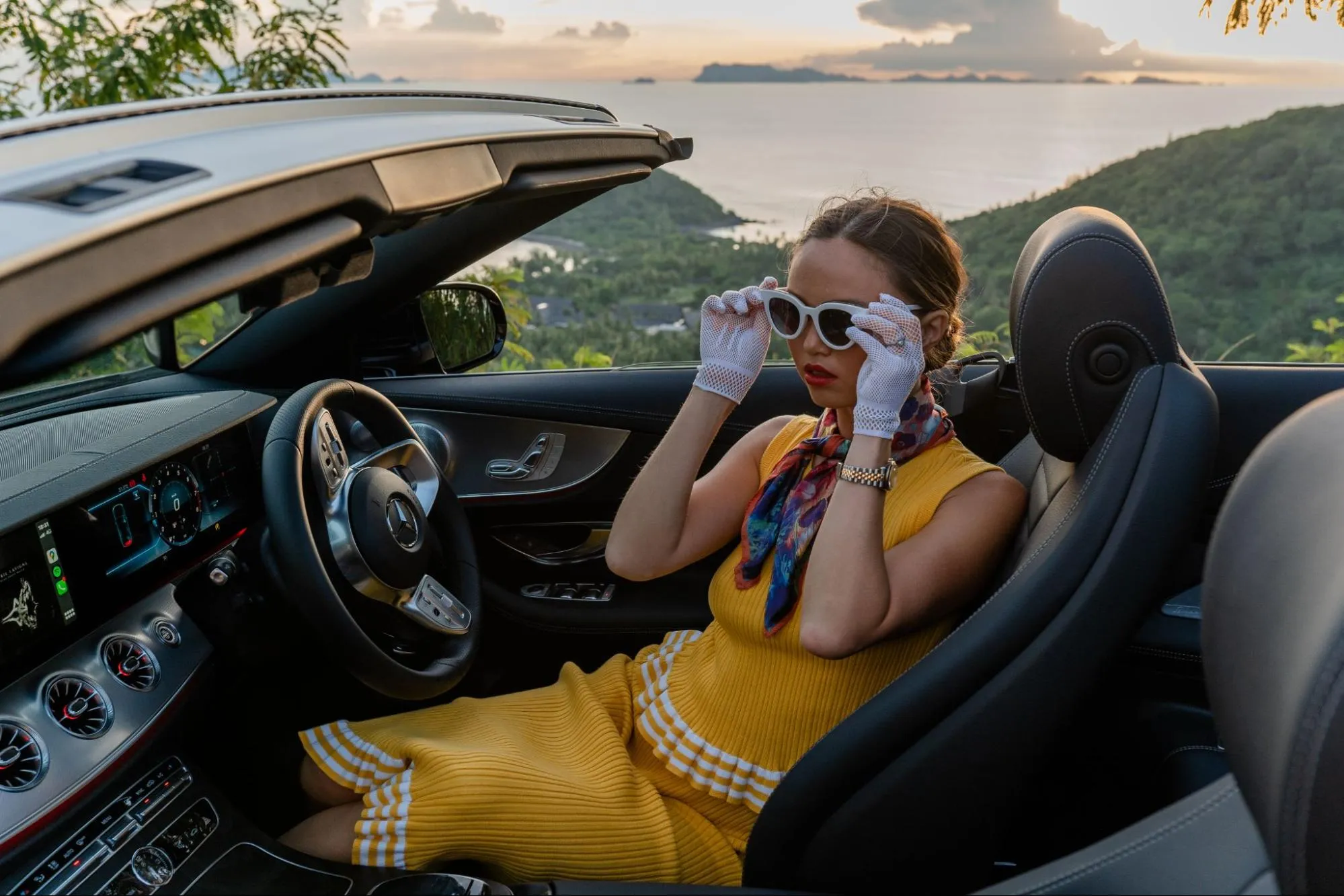 Elegant woman in retro style outfit sitting in luxury convertible car with scenic ocean view at sunset