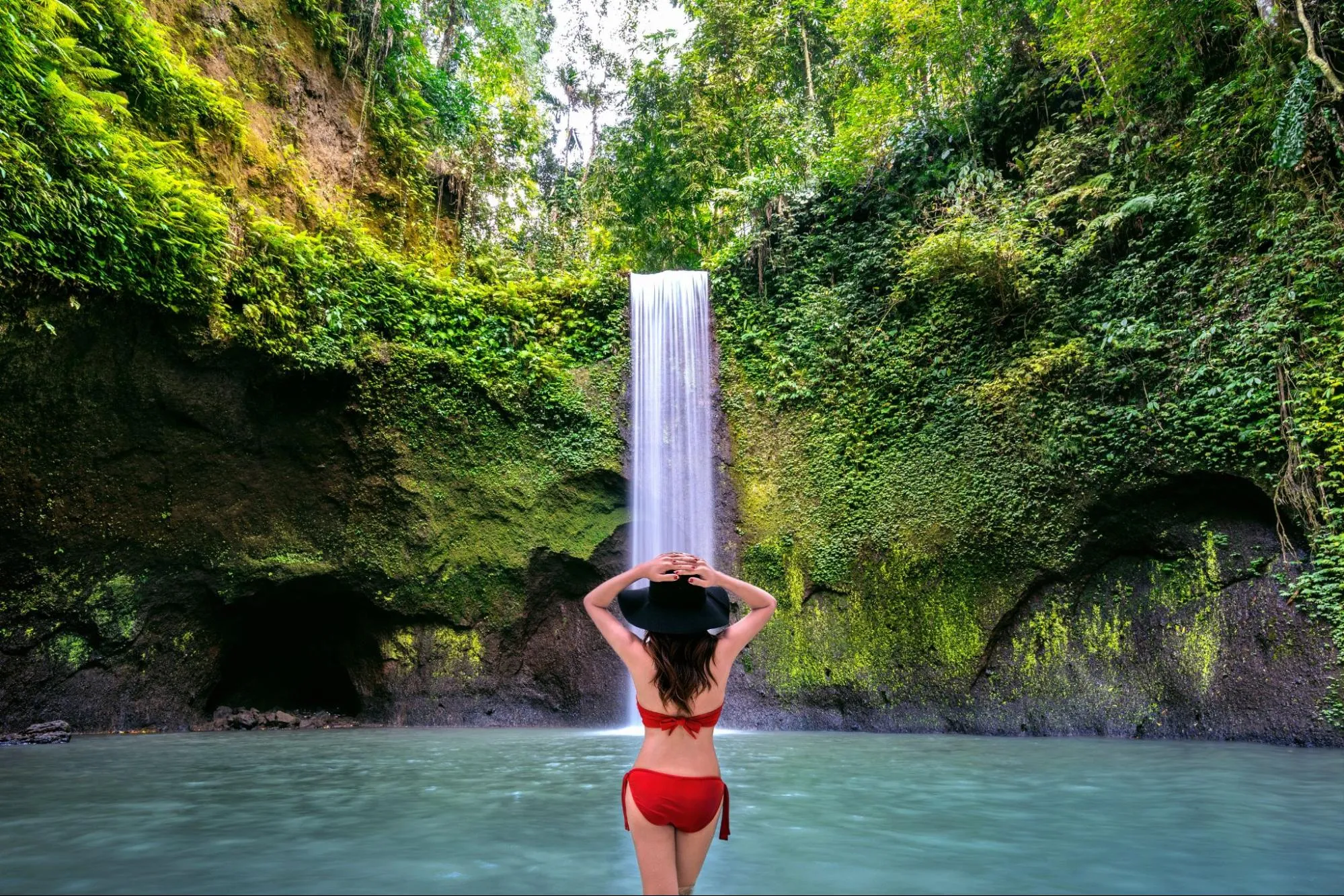 Woman in red bikini standing in front of tropical waterfall surrounded by lush jungle