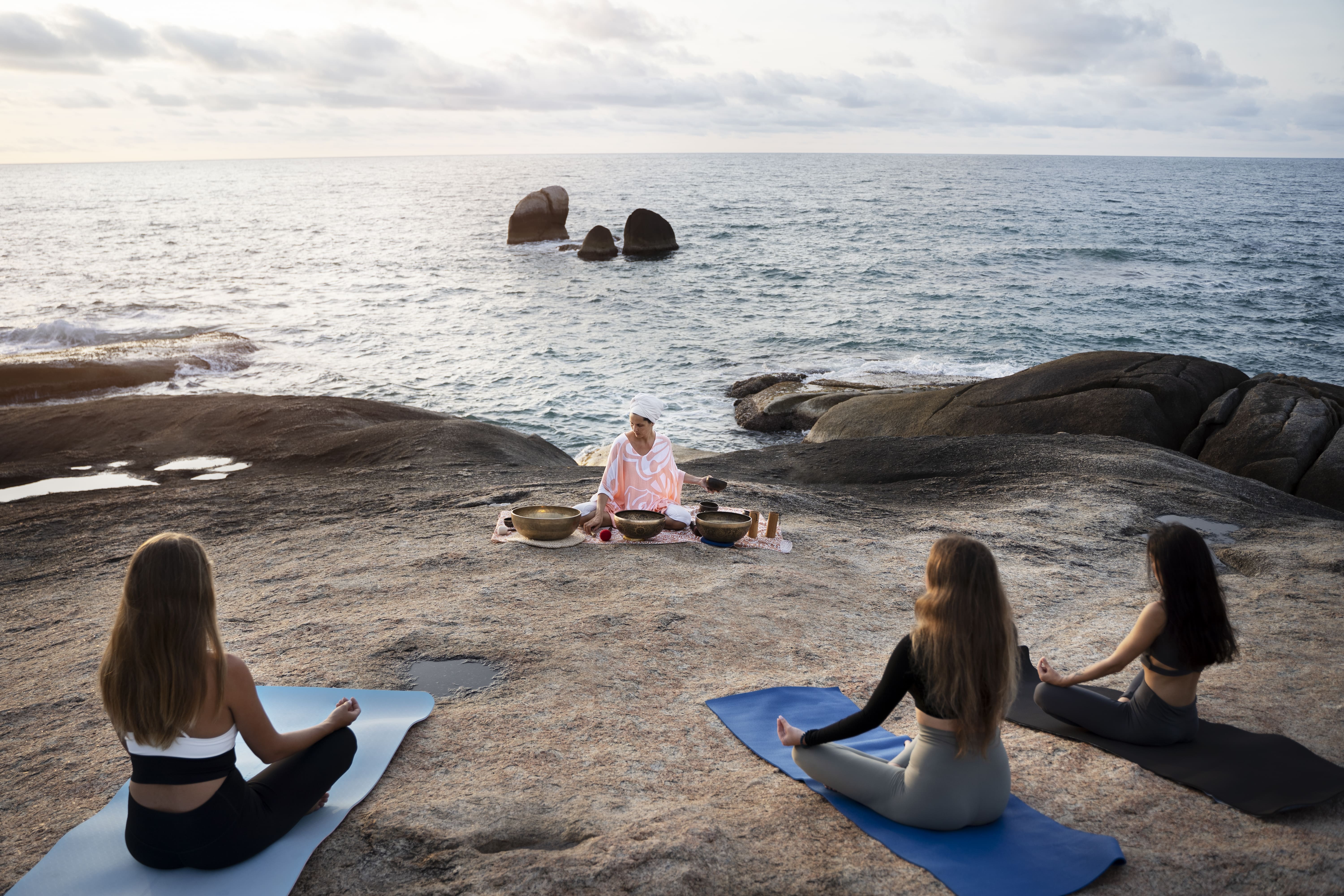 Group of women practicing meditation and sound healing with singing bowls on rocks by the ocean in the Dominican Republic.