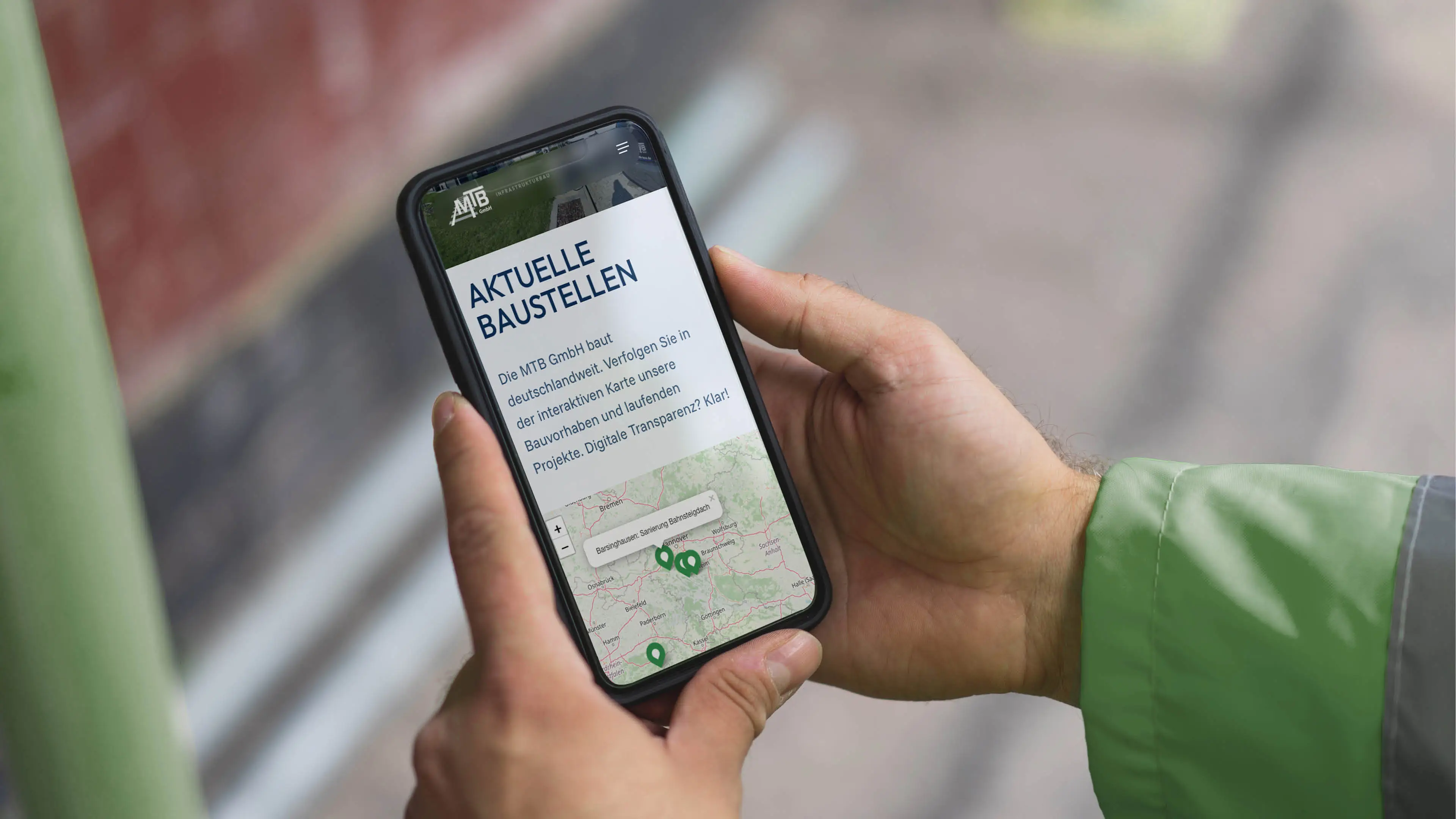 Foto von einem Gleisbauarbeiter der ein Handy in der Hand hält auf dem die Websiteunterseite "Baustellen" abgebildet ist