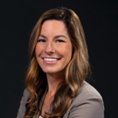 Smiling woman with long wavy hair in a gray blazer against a black background.