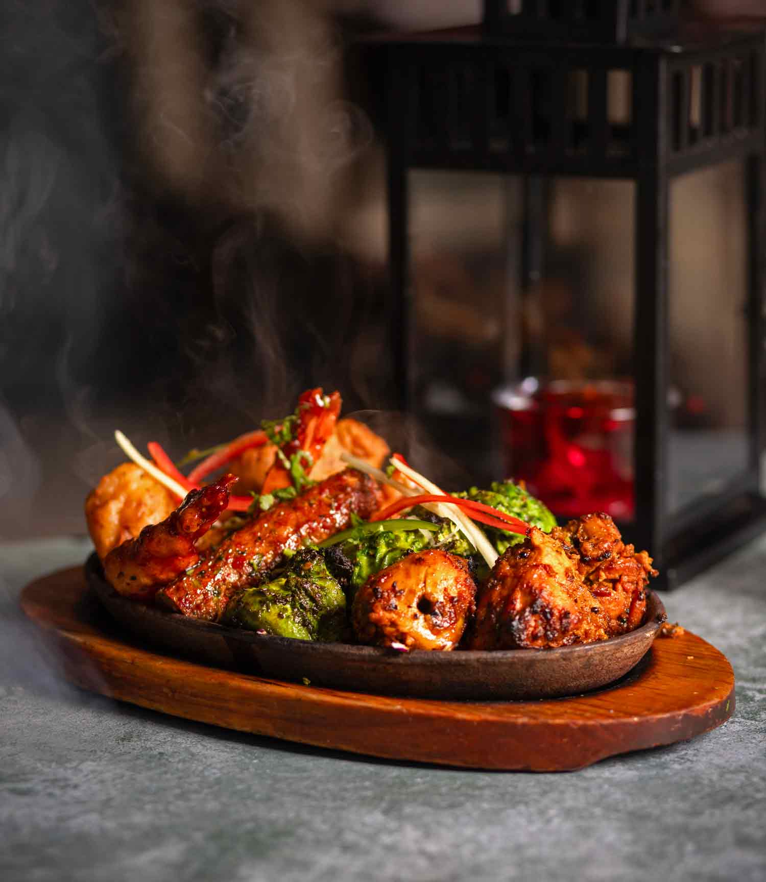 A cast iron plate with a mixed grill on it. There's red chicken, green chicken, sheesh kebabs and more. All these items are charred and they have garnish on top with red and green finely sliced peppers.