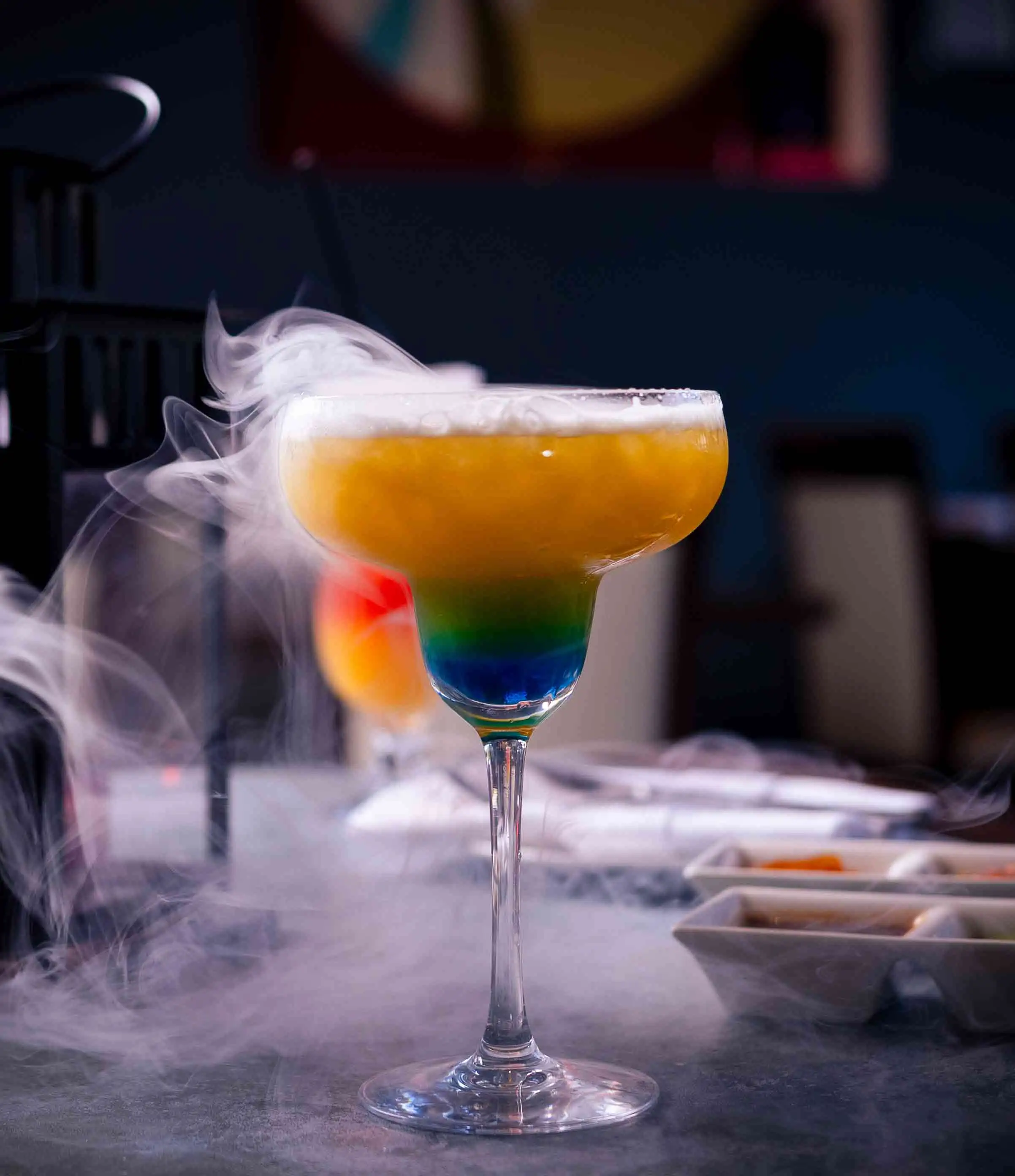 A cocktail in a tall cocktail glass. The cocktail has layers starting with blue, then green and ending with orange. On top of the orange there is white smoke layer and smoke leaving the glass. The glass is on a table where there's food in the background and the food in the background is blurry.