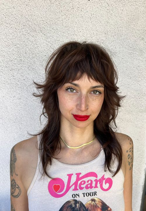 A femme person with a long, shaggy haircut and textured bangs. Their hair is a deep, chestnut brown color with textured pieces framing the face.