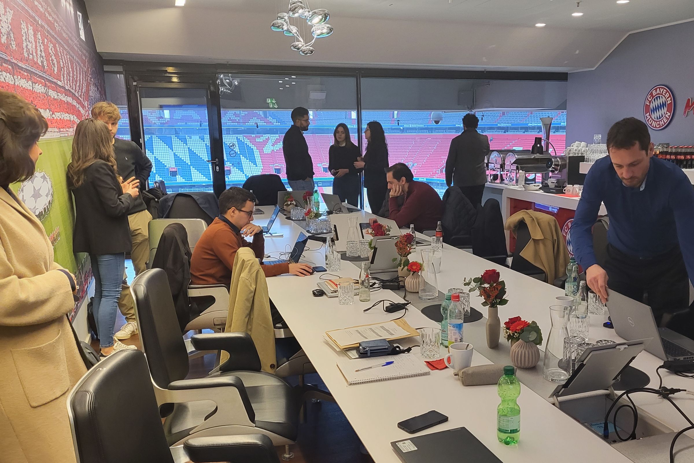 Gemini Projekt Meeting in der Allianz Arena München, zu sehen der lange Tisch, wo Projektleiter an Laptops arbeiten oder sich stehend unterhalten. Ausblick auf die Tribünen vom Stadion mit FC Bayern München Logo