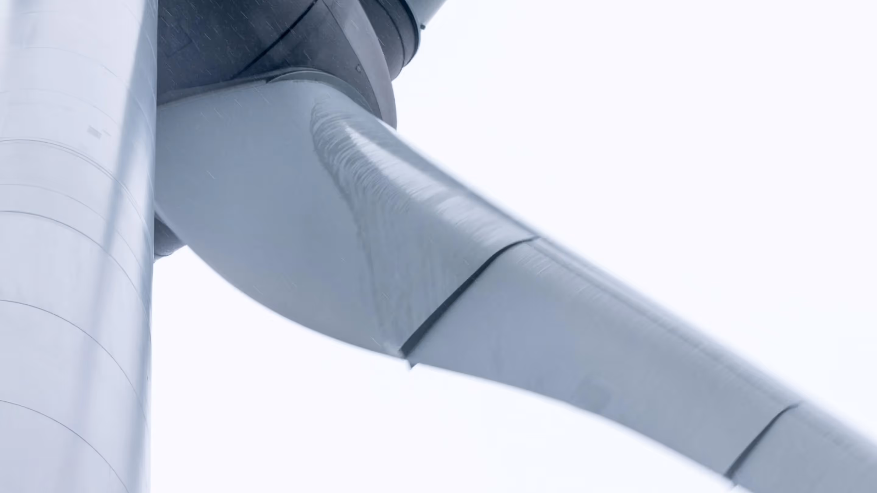 Detail view of a grey wind turbine blade and nacelle with visible rain or mist, against a pale grey sky.