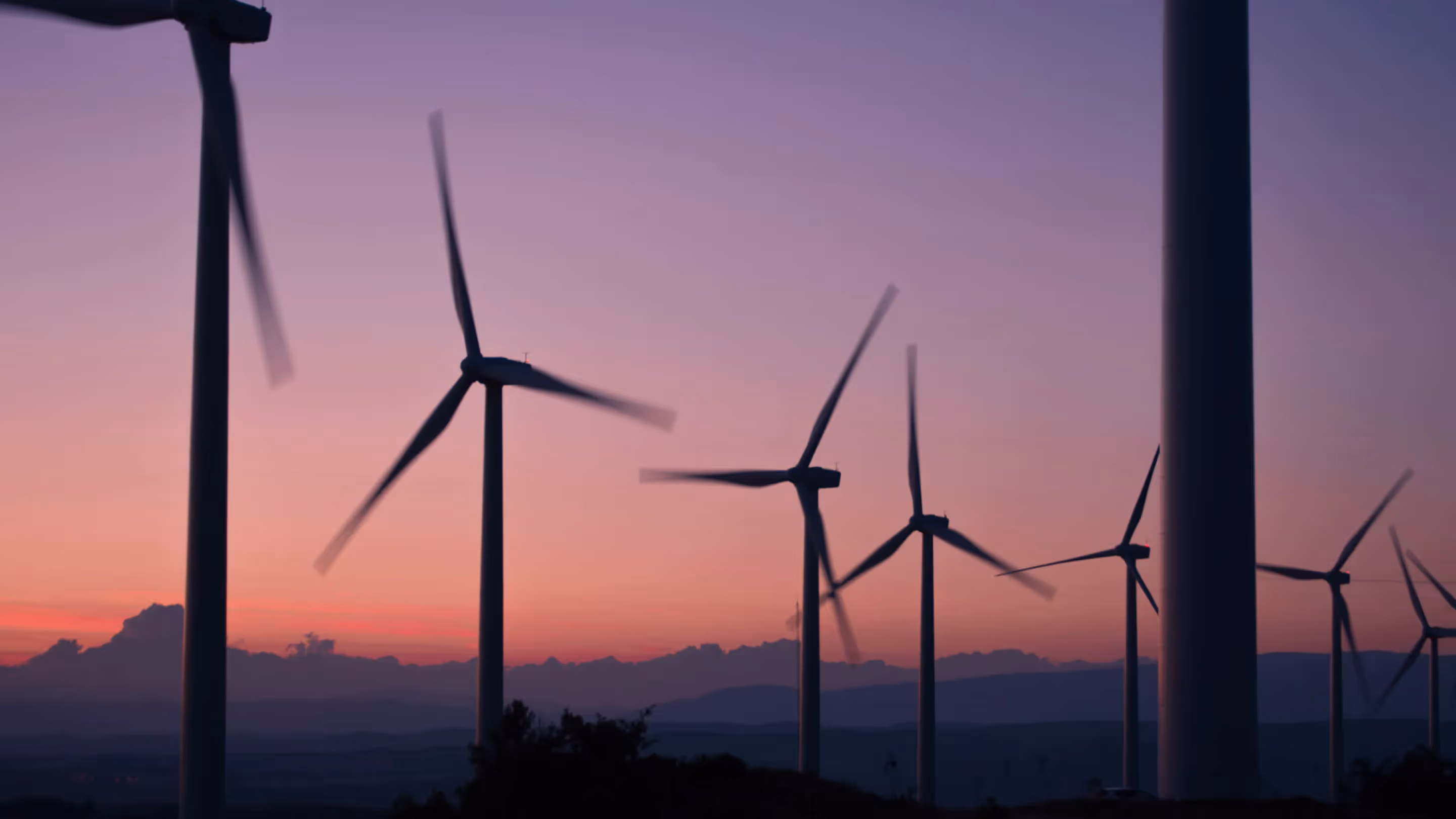 Silhouetted wind turbines spinning against a purple-pink sunset sky, highlighting renewable energy infrastructure and its role in sustainable supply chain development.