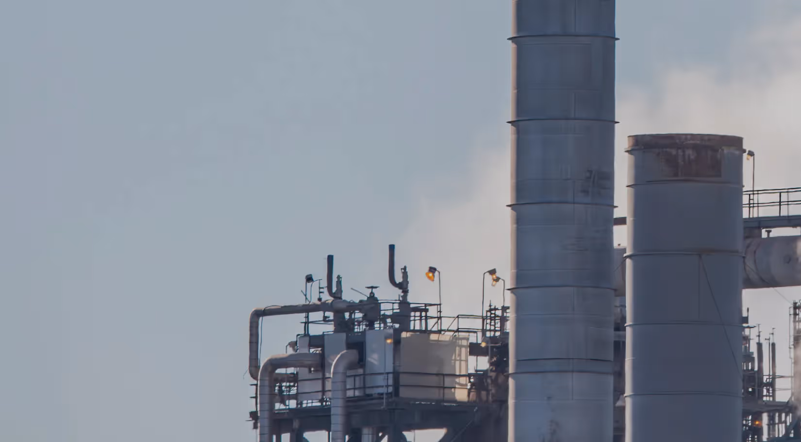 Industrial facility with large metal pipes and smokestacks emitting light smoke against a pale blue sky. The scene highlights heavy industry infrastructure and air emissions.