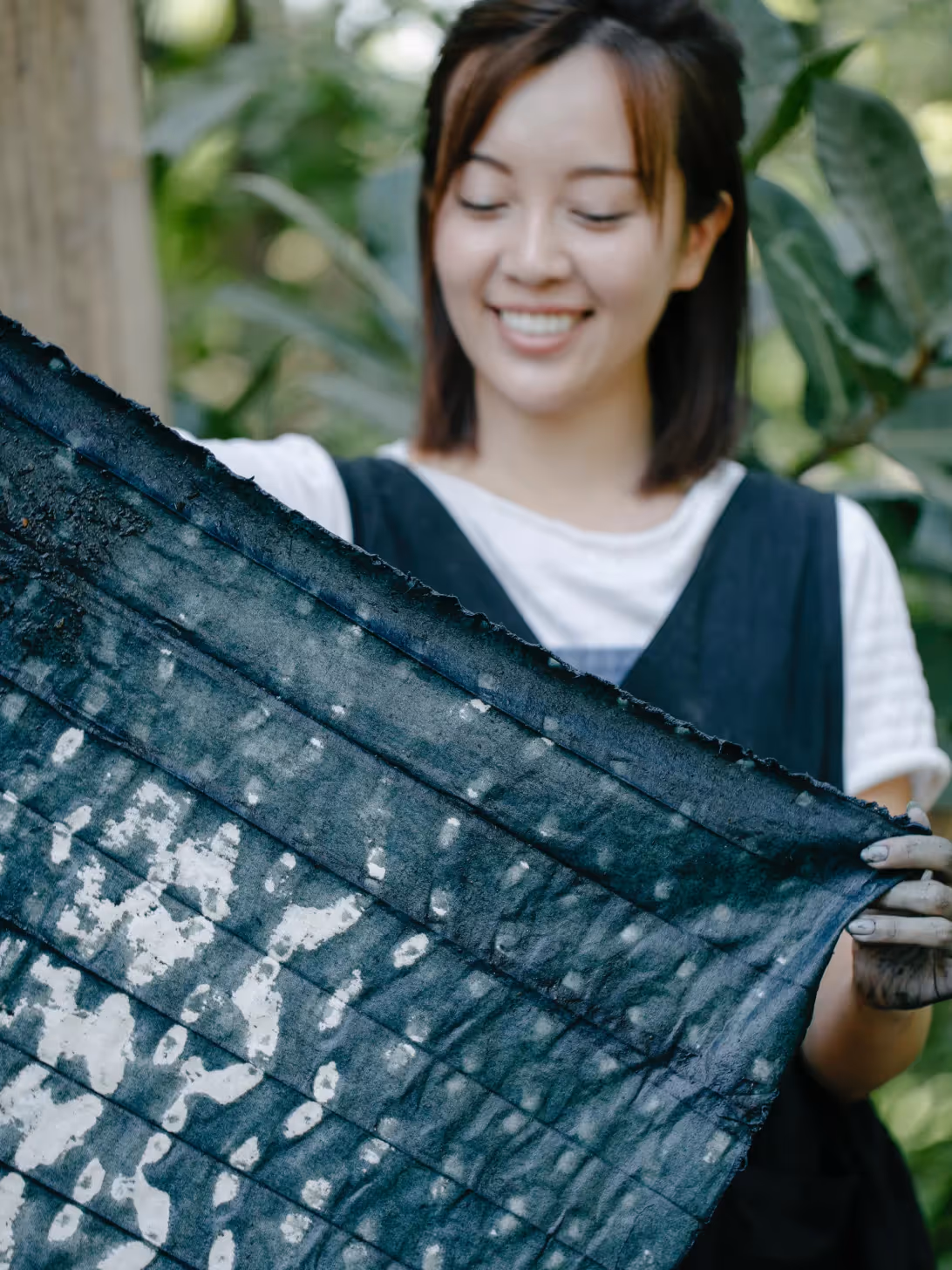 Smiling woman holding up a large piece of hand-dyed indigo fabric outdoors, with visible dye on her hands. She appears engaged in artisanal or small-scale textile production.