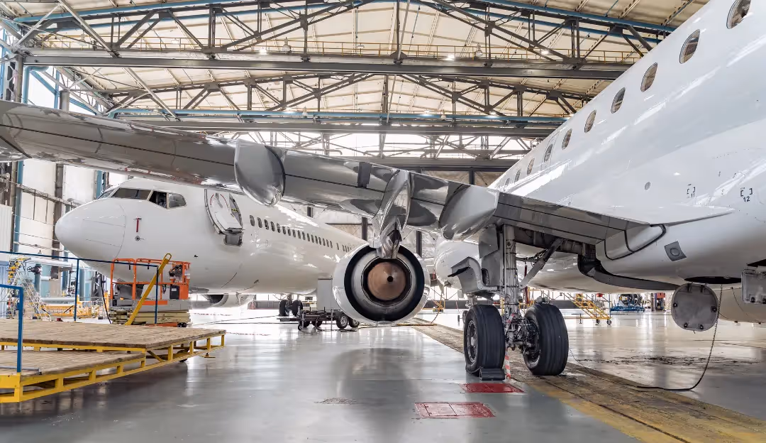 Large cargo ship loaded with multicoloured shipping containers docked at a port during sunset. Several yellow cranes extend over the ship, and the ocean and other vessels are visible in the background.