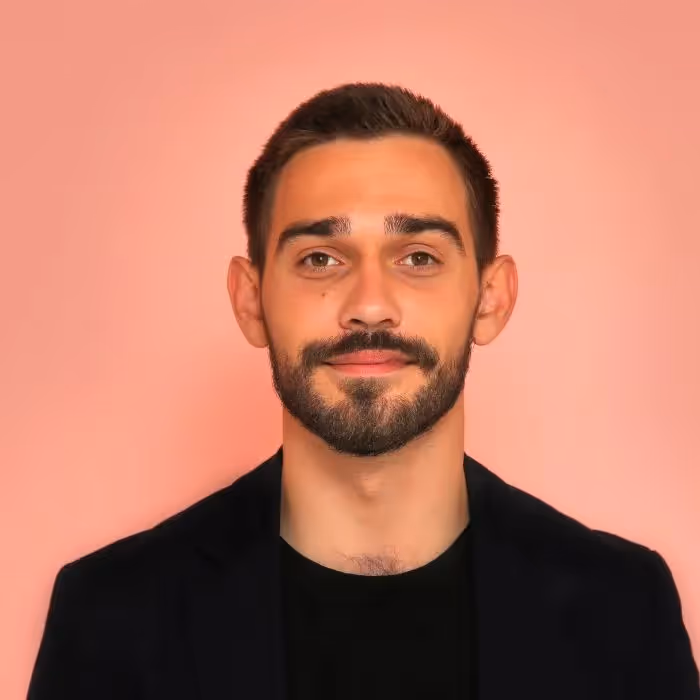 Portrett av en mann med trimmet skjegg, smilende lett, iført en svart skjorte mot en lys rosa bakgrunn.