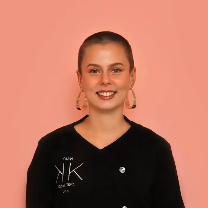 En ung kvinne med et selvsikkert smil, iført en svart skjorte og store ringøredobber, stående mot en vanlig rosa bakgrunn.