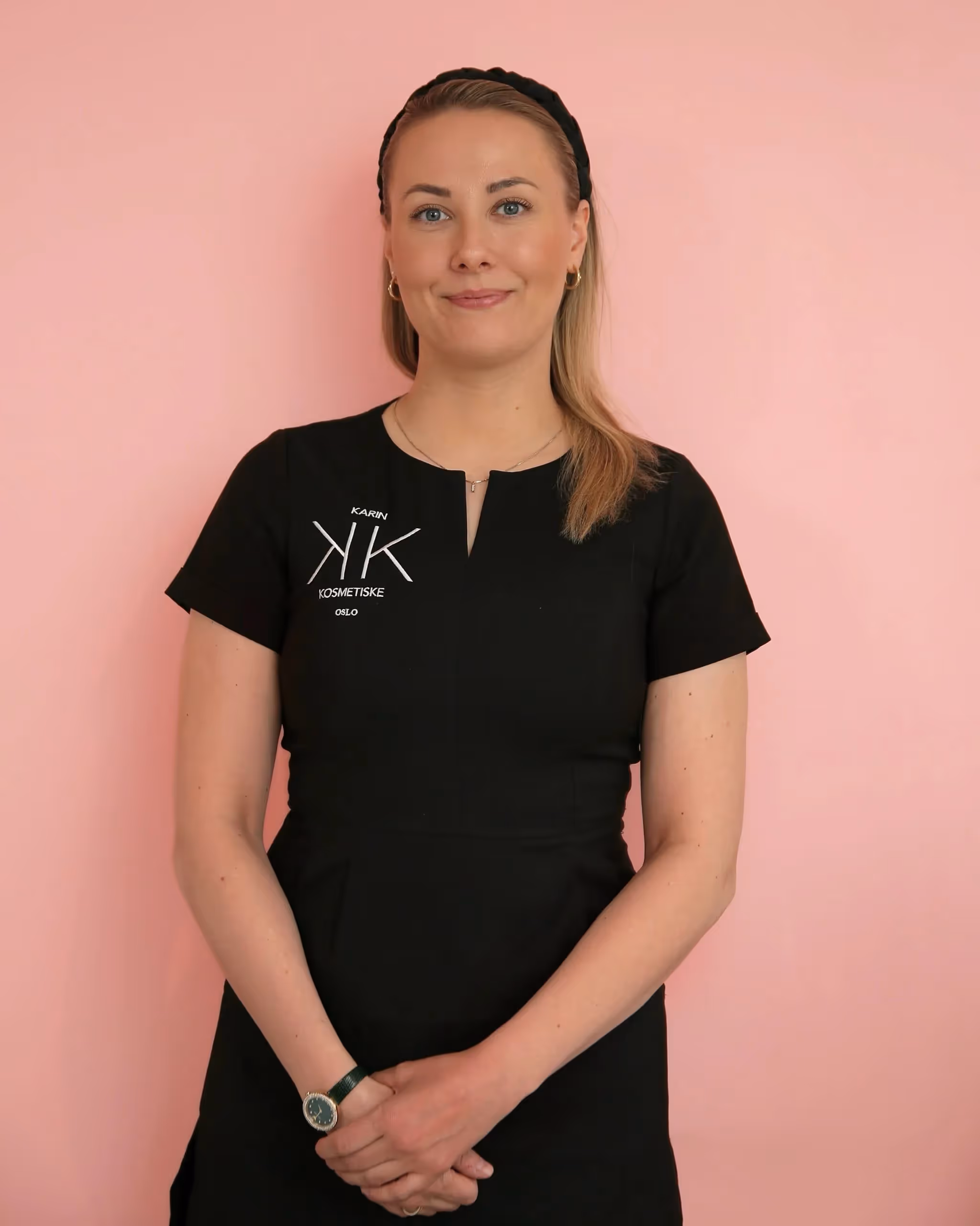 En kvinne med rett, lyst hår iført en svart kortermet kjole med en liten logo på brystet står mot en rosa bakgrunn og smiler lett med hendene knyttet foran seg.